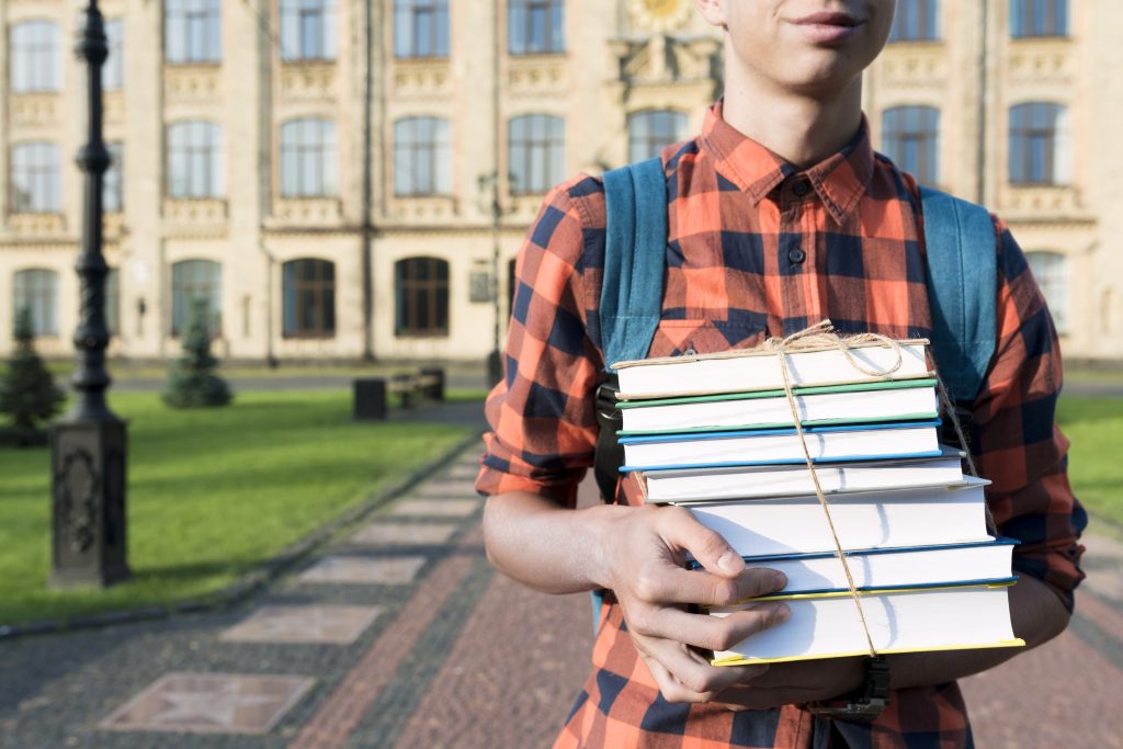 Mężczyzna trzymający stos książek na zewnątrz budynku uniwersytetu.